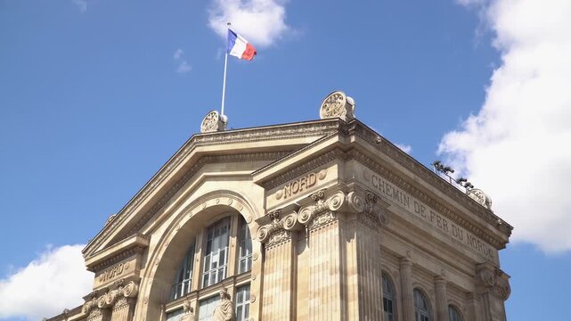French flag waving on Gare du Nord station building in Paris, France. Blue summer sky with white clouds.