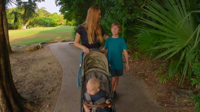 Young mother walking with her two sons in a tropical park, younger baby sitting in a stroller. Warm family walk surrounded by greenery. Slowmotion video.