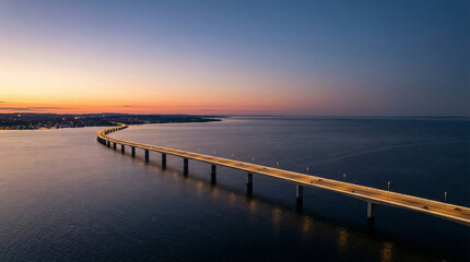 Obraz premium aerial shot of toll road over the sea at dusk with moving cars creating light trails, illuminated lamps, dramatic sunset sky with orange and purple tones, reflections on calm ocean water.