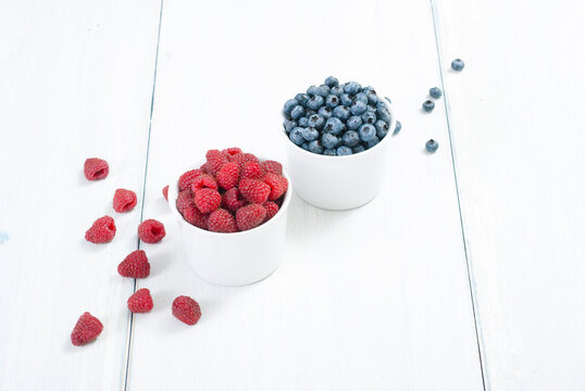 bilberry and raspberry fruits at white ceramic cup, on bright wood table