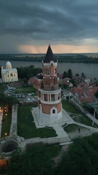 Vertical video drone orbits Gardos Tower at dusk as dramatic storm clouds filled with lightning gather over the Danube river in Zemun, Belgrade, Serbia.