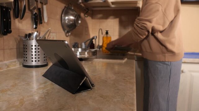 Person doing household chores rinsing dishes in the kitchen sink while viewing content on a digital tablet, blending technology into daily routine in a modern home environment
