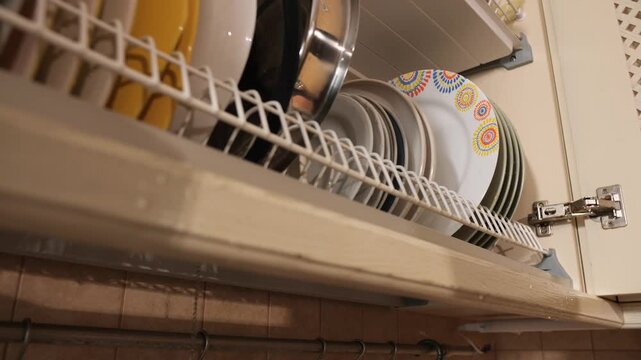 Hand placing a freshly washed, colorful plate onto a drying rack inside an overhead kitchen cabinet, showcasing everyday household chores and organization