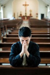 Fototapeta premium Young Asian man praying with clasped hands in a church pew. Christian faith and spirituality concept symbolizing devotion, repentance and peaceful worship inside a cathedral.