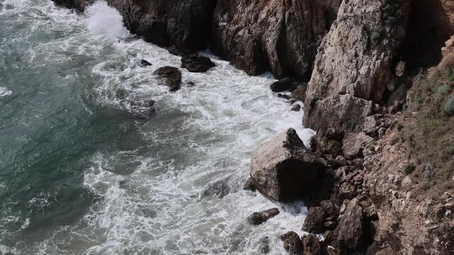 A dynamic top-down shot of white sea foam and powerful waves crashing against the dark rocky cliffs of Sperlonga. The image evokes the raw energy of the Mediterranean Sea along the Riviera di Ulisse. 