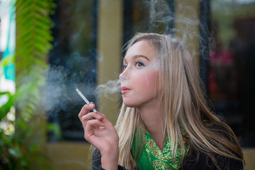 Contemplative Young Woman Smoking White Cigarette Outdoors in Sunny Setting, Wearing Vibrant Patterned Top, Relaxing in Natural Light, Holding White Cigarette in Hand, Sunlight, Patterned Clothing © AU USAnakul