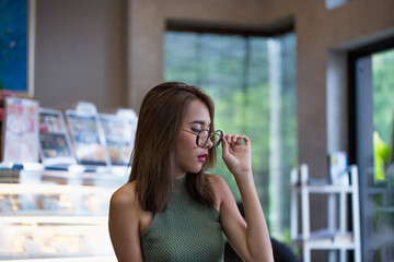 Fototapeta premium Young woman with dark hair and glasses enjoys quiet moment in cozy cafe, engrossed in magazine. She?s seated at table with display case visible in background. warm lighting