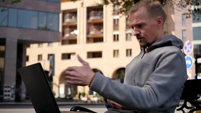 Young man with stylish beard and haircut using laptop while sitting on bench in city and working online outdoors. Person using video call on laptop. Remote work online meeting