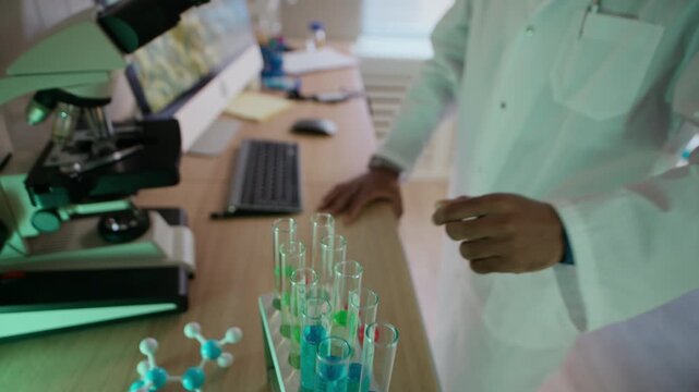 Tilt down shot of male African American researcher in white coat taking empty test tube while examining specimen under microscope in laboratory
