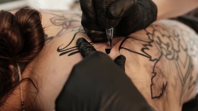 Close-up of a female tattoo artist in black latex gloves using a wireless rotary tattoo pen to ink a large, intricate floral and mandala design on a woman's upper back.