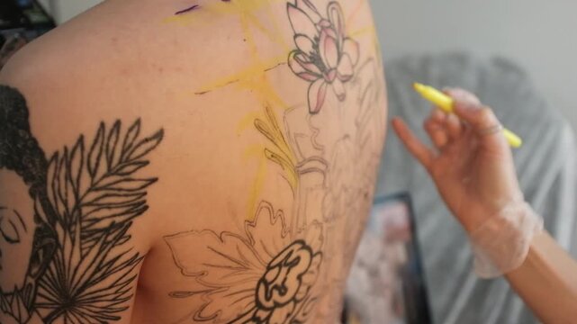 Close-up of a female tattoo artist draws a scketch of intricate floral and mandala design on a woman's upper back. Free hand tattoo technique.
