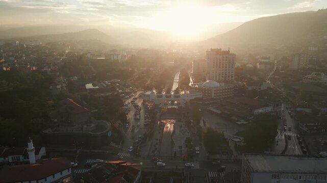Forward drone shot captures Novi Pazar in silhouette against a warm glowing sunset sky, Serbia, Balkans.