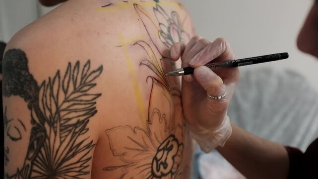 Close-up of a female tattoo artist draws a scketch of intricate floral and mandala design on a woman's upper back. Free hand tattoo technique.