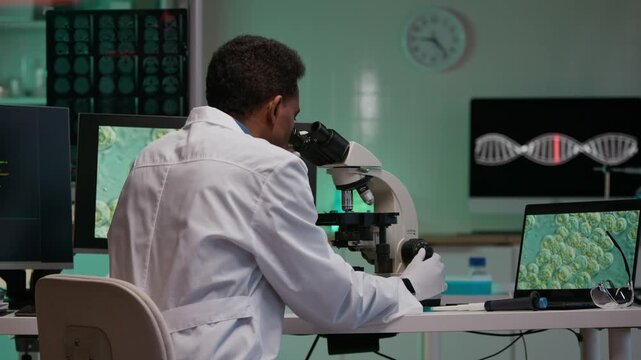 Back view of Black male geneticist analyzing cell morphology and genomic data through microscope and using computers with digital software in smart laboratory