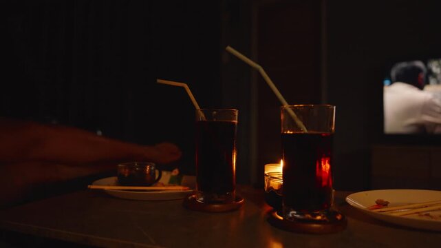 Dimly Lit Hands Lifting Tall Glasses of Dark Drinks with Candles Flickering Beside on Table side low angle Cozy Leisure Drinks Concept