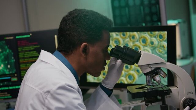 Side view of young male molecular biologist evaluating mutation of cells through microscope in modern laboratory with artificial intelligence applications on computers
