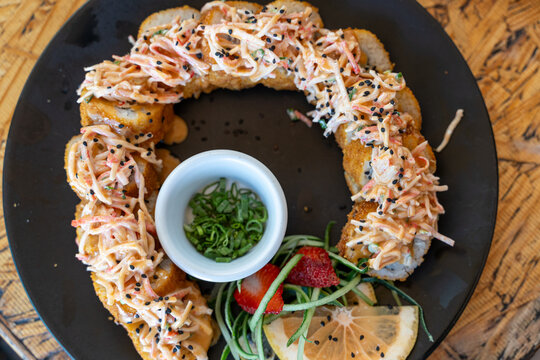 Top view of crispy fried sushi roll topped with creamy spicy mixture, served with dipping sauce, lemon slices, and fresh vegetables