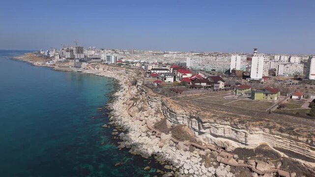 Stunning aerial drone footage of the Aktau coastline on a bright summer day. The shot captures a panoramic view of the Caspian Sea shore, featuring a mix of modern high-rise buildings, private cottage