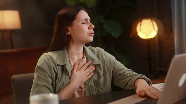 Worried young woman working on laptop while has panic heart attack hold hand on chest and try to calm down alone Anxious female with mental health problem at home