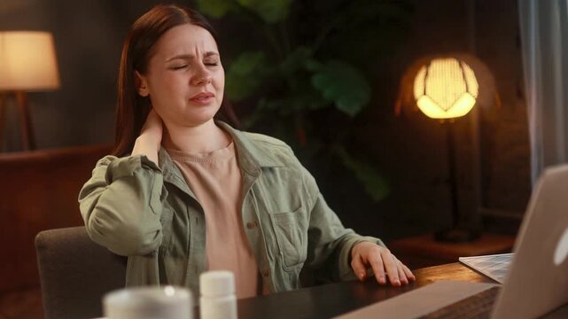 White Woman Working At Home Rubbing Back And Neck. Freelance Writer Pauses At Late Evening Desk, Massaging Lower Back And Stretching Near Laptop And Lamp, Pills On Table, Shows Tension,