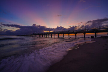 Obraz premium Sunset light spreads across the sky as a long pier stretches into the sea, with gentle waves reflecting purple and gold tones along the shore.