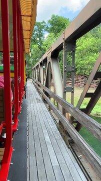 A moving theme park train crosses a rustic wooden bridge above calm green water surrounded by trees. Metal beams and natural scenery create a peaceful travel and exploration mood.