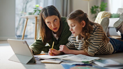 Obraz premium Relaxed babysitter teaching girl online homework on tablet lying floor closeup