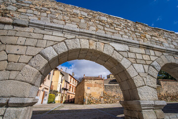 Naklejka premium A historic stone archway of the ancient Segovia Aqueduct in Spain, framing a charming street with traditional buildings under a bright blue sky. This architectural marvel dates back over 2000 years.