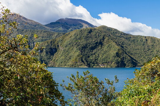 A  panoramic view of Laguna de Cuicocha, a deep crater lake located within the Cotacachi-Cayapas Ecological Reserve in the Ecuadorian Andes. Two volcanic islands, lush caldera walls under a bright sky