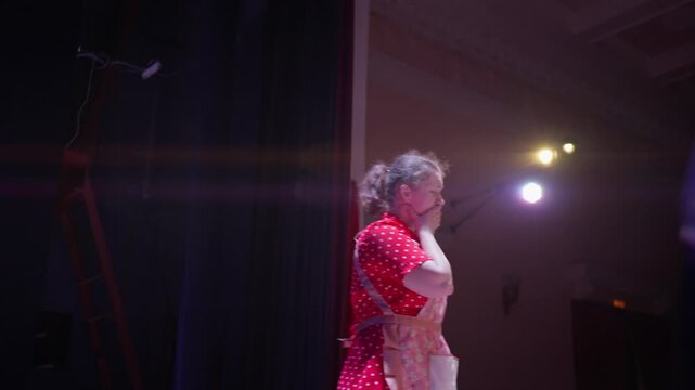 Solo stage performance of a woman in a red polka dot dress illuminated by warm theater lights as she rehearses a dramatic scene in an empty auditorium