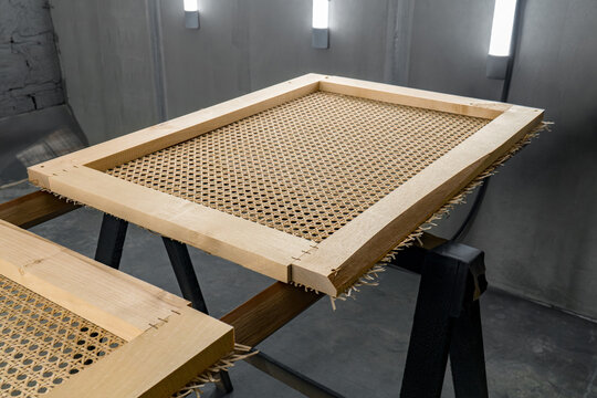 Wooden frame with tightly woven rattan surface sits on a stand in a paint booth. the frame has neat joints, rattan edges slightly frayed. industrial background with bright lights
