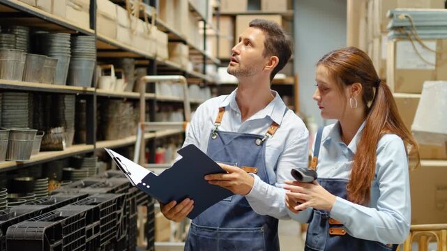 Diverse small business owners in aprons working in a warehouse, checking inventory stock on shelves with a clipboard and barcode scanner. Teamwork in retail logistics and supply chain management.