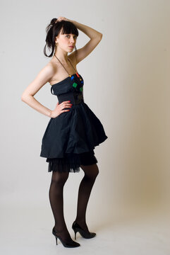 Studio portrait of a young woman wearing an elegant black dress, posing against a clean white background. The model is captured in a full-length or three-quarter pose, exuding confidence