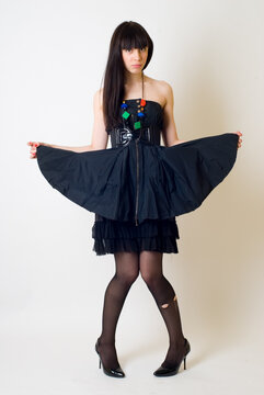 Studio portrait of a young woman wearing an elegant black dress, posing against a clean white background. The model is captured in a full-length or three-quarter pose, exuding confidence