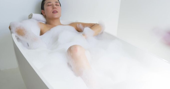 Camera pulling back while woman reclining in bathtub with foam, resting on rolled towel relaxing