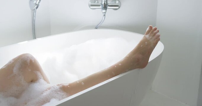 African American woman reclining in bathtub, shifting in foam, extending legs and rinsing by faucet
