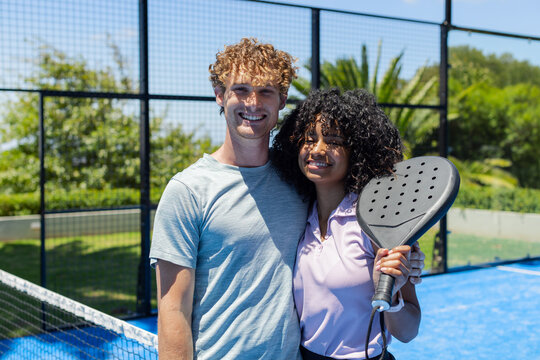 Couple standing in sportswear on blue padel court near net and fence, woman holding black racket