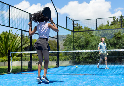 Female and male players jumping and watching ball on blue fenced padel court, net and paddles