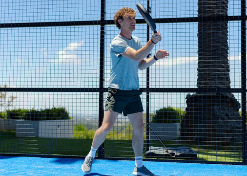 Man wearing light blue tee holding paddle racket and watching incoming shot on blue padel court