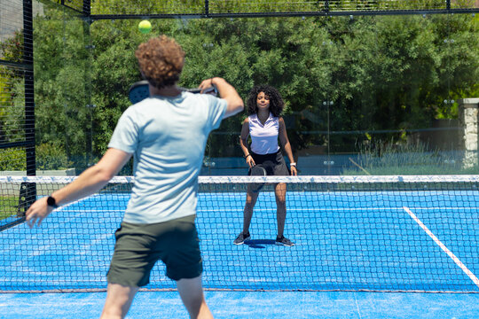 Male and female players swinging paddles, preparing to return ball crossing net on blue padel court