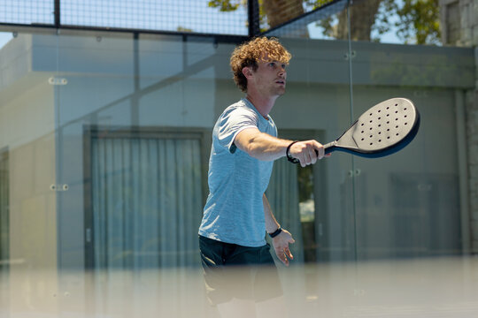 Adult male athlete standing inside glass paddle court holding padel racket and looking upward