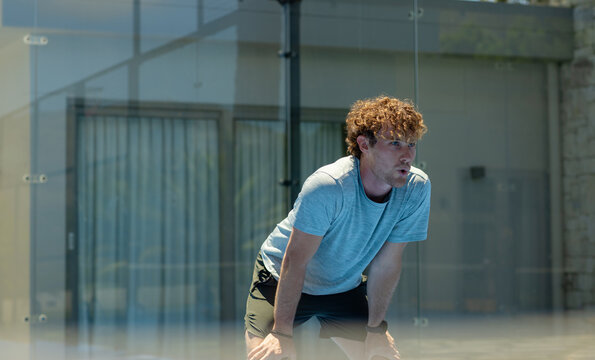 Adult man bending, breathing heavily on glass court by metal post, wearing athletic gear and watch