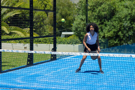 Adult female in polo and shorts standing on blue padel court holding paddle returning yellow ball