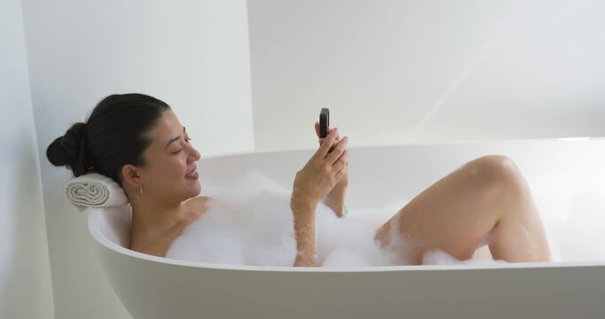 African American woman relaxing on rolled towel, reclining in white tub amid bubbles, tapping phone
