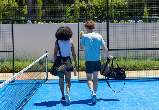 Diverse friends walking away across blue paddle court carrying sports bags, rackets, ball, bottle