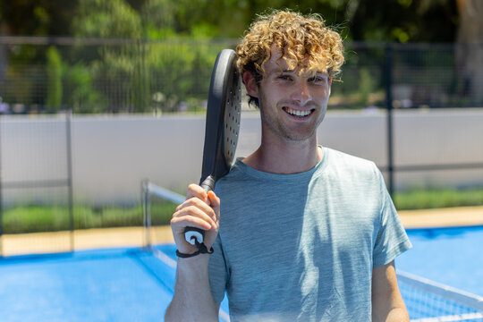 Male standing on blue court with net holding black perforated racket, wearing light-blue shirt