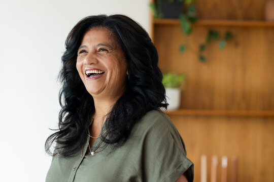 Mature African American woman laughing, looking left in room with wood shelf, plants, olive blouse