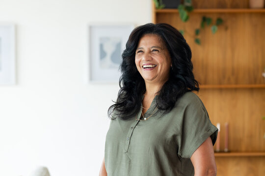 Mature African American woman standing laughing at home in olive green blouse and layered necklaces