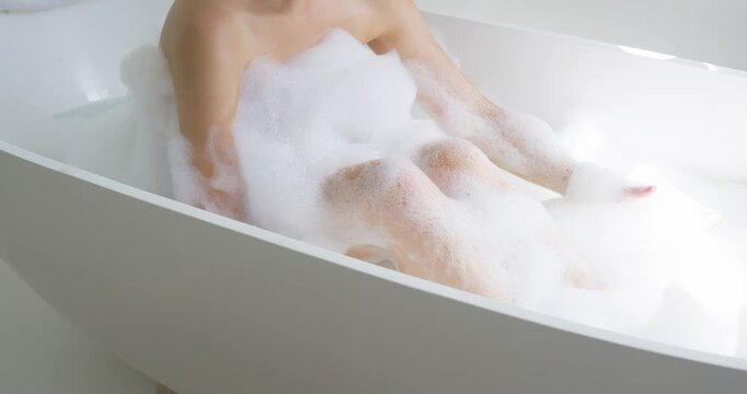 Adult woman is shifting arms and compressing bath foam in white freestanding bathtub for comfort
