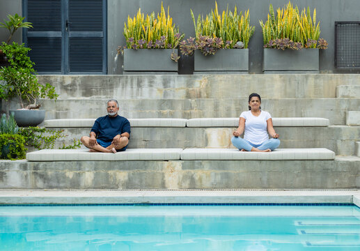 African American senior couple sitting cross-legged on cushioned benches by pool with planters